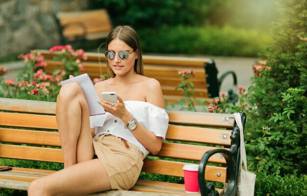 Jolie jeune femme en vêtements d'été et lunettes de soleil tenant smartphon