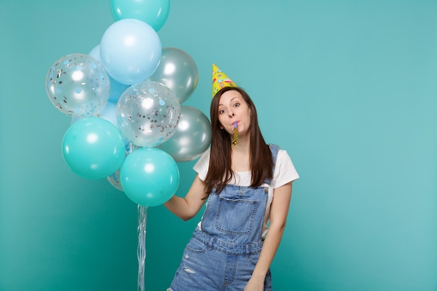 Jolie jeune femme en vêtements en jean, chapeau d'anniversaire soufflant dans un tuyau, célébrant et tenant des ballons à air colorés isolés sur fond bleu turquoise. Fête d'anniversaire, concept d'émotions de personnes.