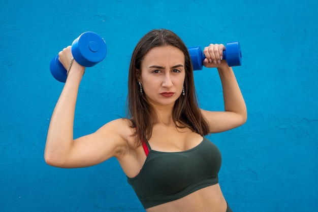 Jolie jeune femme en vêtements de sport verts tient des haltères dans ses mains sur fond bleu. Mode de vie sain