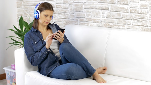 Jolie jeune femme vêtue de jeans écouter de la musique avec des écouteurs sur un canapé blanc à la maison