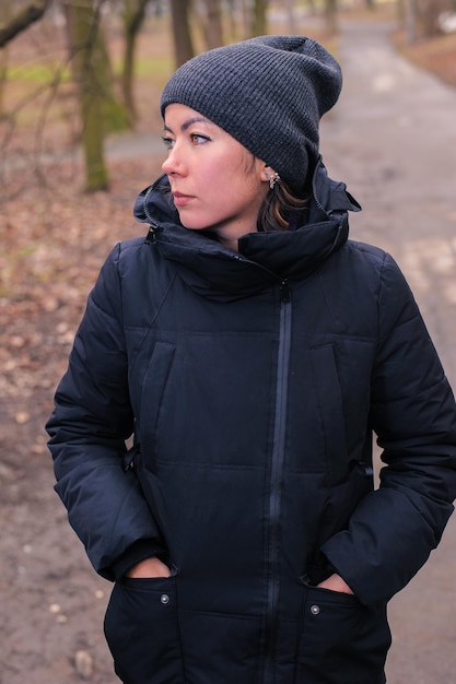 Une jolie jeune femme vêtue de vêtements d'hiver noirs modernes, d'un manteau et d'un chapeau tricoté se promène dans le parc.