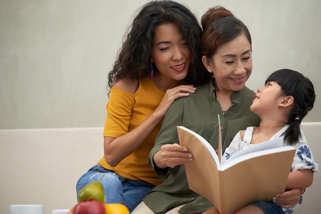 Jolie jeune femme vietnamienne aimante embrassant une mère âgée pendant qu'elle lit un livre à sa petite-fille