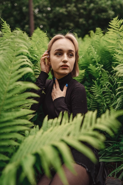 Jolie jeune fille aux cheveux blonds courts est assise sur le sol parmi les fougères dans un pré d'été