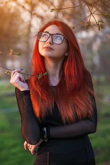 Jolie jeune fille aux cheveux rouges, portant des lunettes, posant près d'un arbre