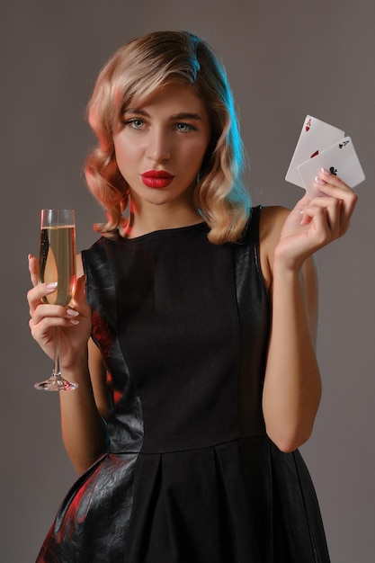 Jolie jeune fille bouclée blonde maquillage lumineux en robe noire élégante tenant deux cartes à jouer et un verre de champagne posant sur fond de studio gris Gambling poker casino Closeup