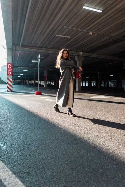 Jolie jeune fille bouclée dans des vêtements de ville à la mode avec un long manteau et un sac à main élégant se promène dans la ville au parking