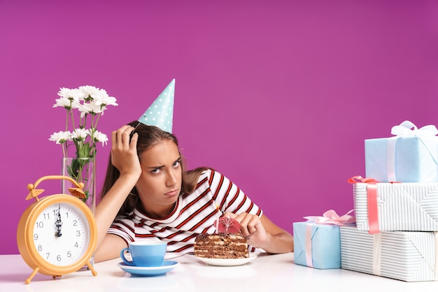 Jolie jeune fille bouleversée assise au bureau avec un gâteau d'anniversaire isolé sur un mur rose