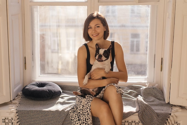 Jolie jeune fille brune caucasienne est assise sur le rebord de la fenêtre avec son bouledogue de compagnie Amour entre chien et concept de propriétaire