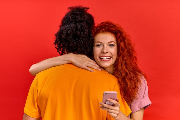 Jolie jeune fille caucasienne aux cheveux rouges heureuse embrasse son petit ami afro-américain et regarde un téléphone intelligent moderne. Concept de site de rencontre en ligne et d'applications pour les amoureux. Espace publicitaire