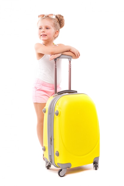 Jolie jeune fille en chemise blanche, short rose et lunettes de soleil se tiennent près de la valise jaune