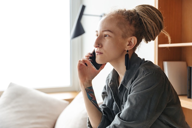 jolie jeune fille concentrée avec des dreadlocks et piercing à l'intérieur près de l'étagère parlant par téléphone portable.