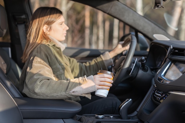 Jolie jeune fille conduisant une voiture d'une main et buvant du café Femme néglige la sécurité dans le véhicule