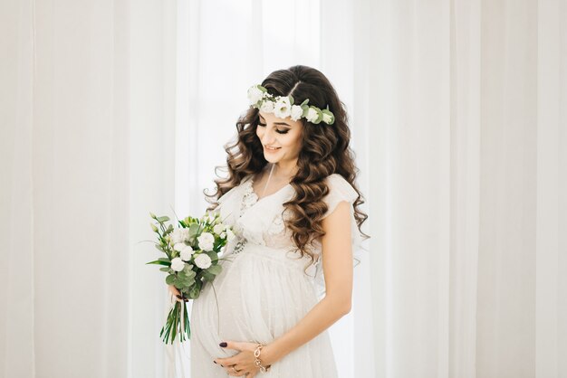 Jolie jeune fille enceinte dans une robe blanche avec des boucles, une couronne et un bouquet de fleurs.