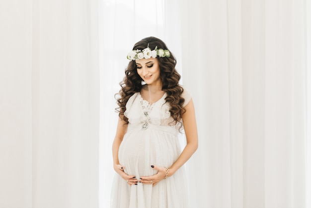 Jolie jeune fille enceinte dans une robe blanche avec des boucles, une couronne et un bouquet de fleurs.