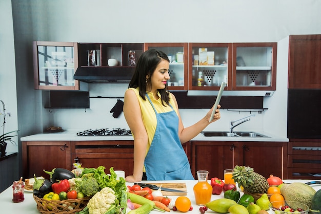 Jolie jeune fille ou femme indienne avec tablier en cuisine multitâche, à l'aide d'un smartphone, d'une tablette avec une table pleine de fruits et légumes et d'un ordinateur