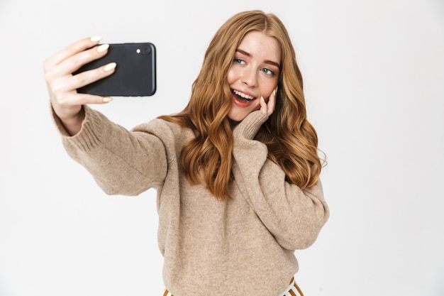 Jolie jeune fille heureuse portant un pull debout isolé sur un mur blanc, prenant un selfie