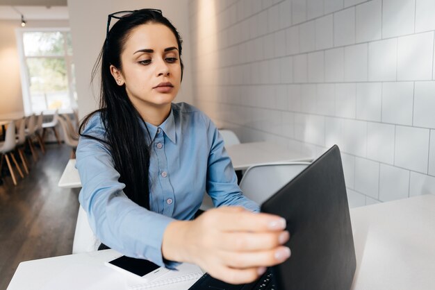 Jolie jeune fille indépendante en chemise bleue travaillant sur ordinateur portable au café