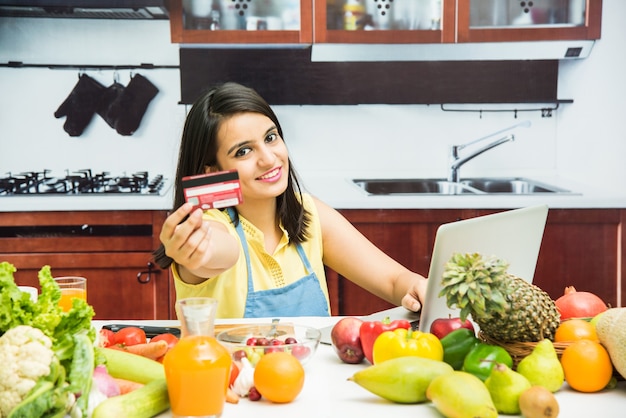 Jolie jeune fille indienne avec tablier dans la cuisine, achats en ligne avec carte de débit ou de crédit sur ordinateur portable avec table pleine de fruits et légumes frais