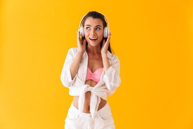 Jolie jeune fille joyeuse portant des vêtements de plage d'été dansant tout en écoutant de la musique avec des écouteurs isolés sur un mur jaune