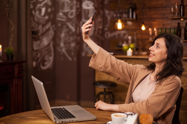 Jolie jeune fille parlant un selfie avec son téléphone portable dans un café moderne. Jeune femme se relaxant dans une cafétéria pendant les pauses. Femme avec ordinateur portable devant elle.