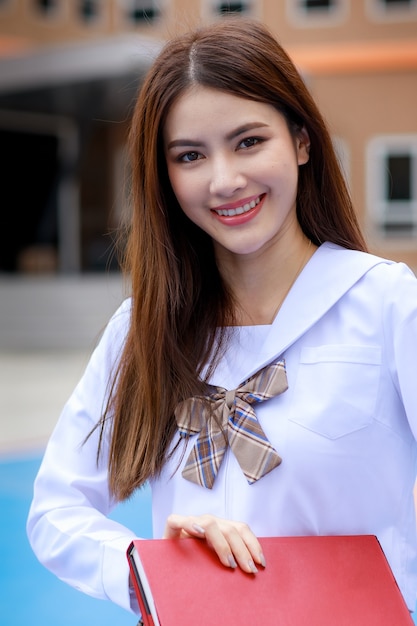 Jolie et jeune fille portant un uniforme d'écolière de style japonais et coréen tenant un livre et pose devant la caméra avec plaisir et plaisir devant le bâtiment de l'école.