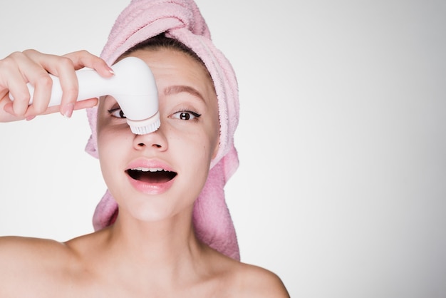 Jolie jeune fille avec une serviette rose sur la tête faisant un nettoyage en profondeur de la peau du visage avec une brosse électrique
