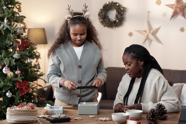 Jolie jeune fille avec un smartphone photographiant un cadeau de Noël fait à la main dans une boîte-cadeau tout en emballant des cadeaux pour des amis