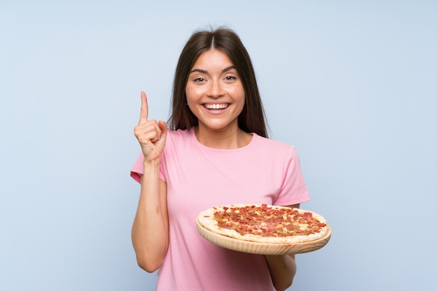 Jolie jeune fille tenant une pizza sur un mur bleu isolé pointant vers le haut une excellente idée