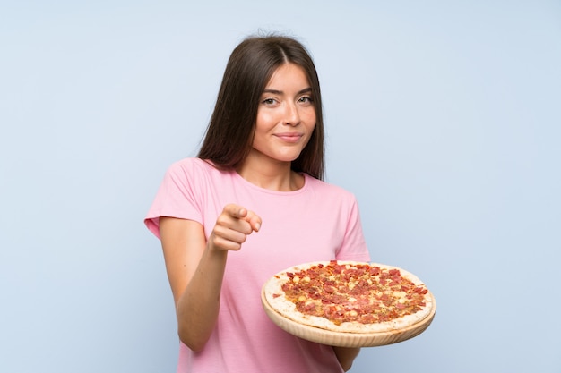 Jolie jeune fille tenant une pizza sur un mur bleu isolé pointe le doigt vers vous avec une expression confiante