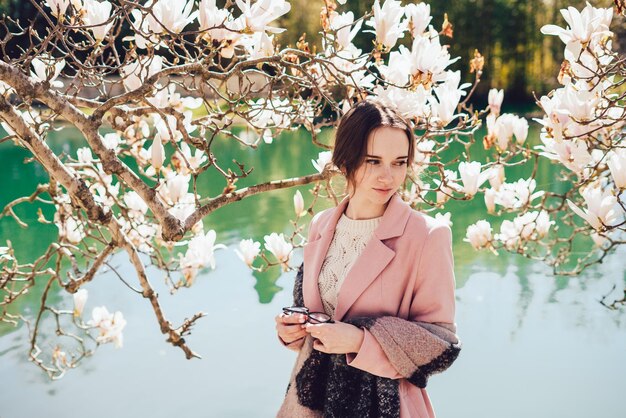 Jolie jeune fille en veste rose posant dans le parc à côté du magnolia en fleurs et de l'étang