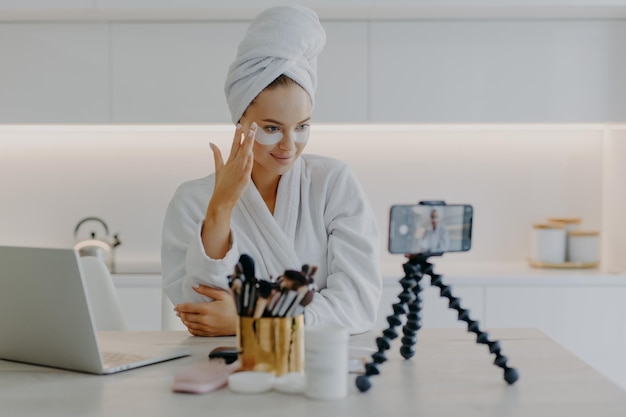 Une jolie jeune mannequin fait de la publicité pour des vues sur Internet. Un produit cosmétique se soucie de la peau sous les yeux. Applique des patchs d'hydrogel. Enregistre une vidéo sur un smartphone pour son blog. Concept de vlogging.