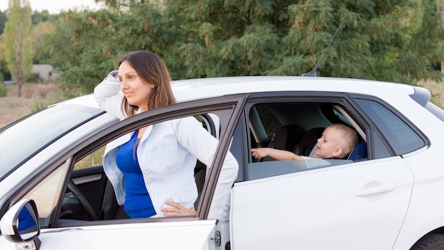 Une jolie jeune mère attend dans la porte de son véhicule garé avec son fils à l'arrière de la voiture