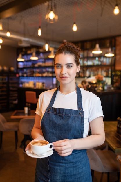 Jolie jeune serveuse brune avec une tasse de cappuccino debout devant la caméra sur fond de tables et chaises au café