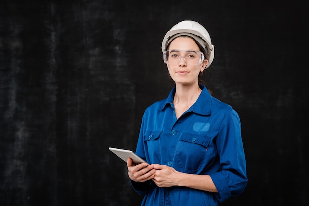 Jolie jeune spécialiste du casque et des vêtements de travail bleus à l'aide du pavé tactile tout en se tenant devant la caméra en isolement