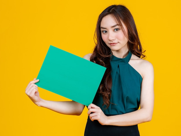 Jolie et jolie brune aux cheveux bouclés tenant un tableau vierge vert pose à la caméra avec un geste joyeux et positif à des fins publicitaires, prise de vue en studio isolée sur fond jaune.