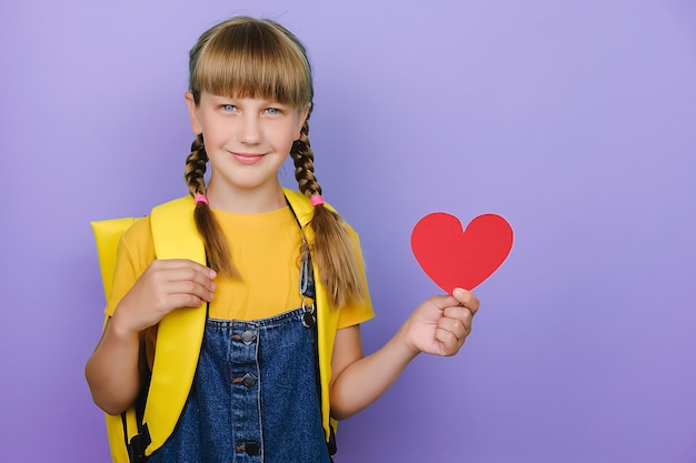 Jolie jolie écolière blonde tenant un petit coeur rouge en papier, portant un sac à dos jaune, isolée sur fond de couleur violet pastel avec espace de copie pour le contenu promotionnel. Concept d'amour et de retour à l'école