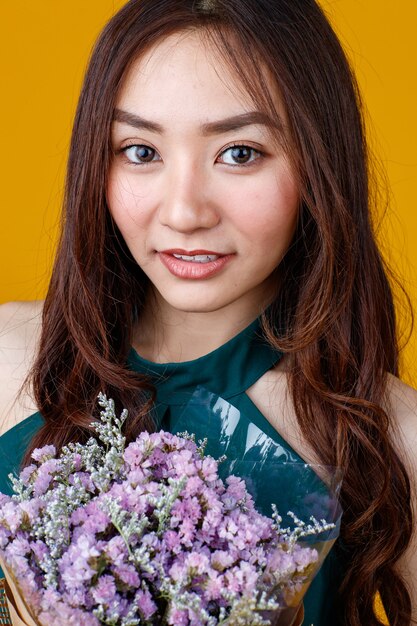 Jolie et jolie femme brune asiatique aux cheveux bouclés tenant un bouquet de fleurs avec gai et heureux, tourné en studio isolé sur fond jaune vif.