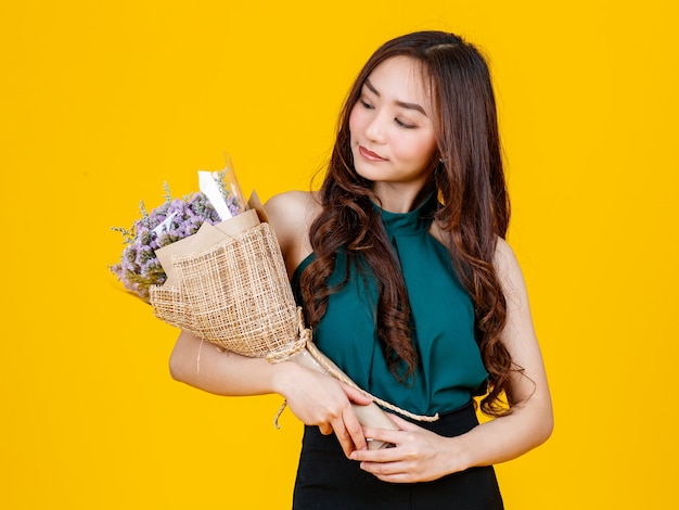 Jolie et jolie femme brune asiatique aux cheveux bouclés tenant un bouquet de fleurs avec gai et heureux, tourné en studio isolé sur fond jaune vif.