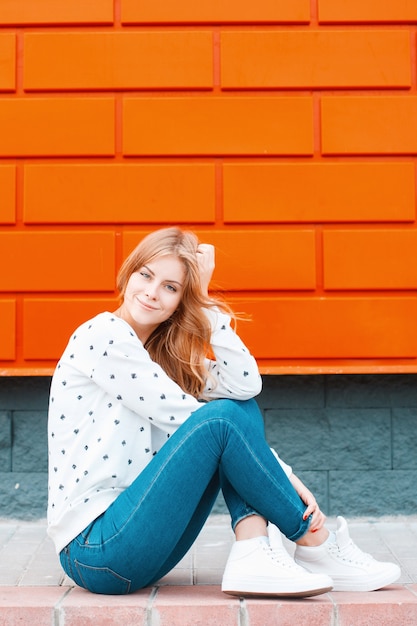 Jolie jolie jeune femme élégante européenne en baskets en jean bleu dans un pull est assis sur le trottoir près d'un bâtiment noir et orange par une chaude journée de printemps