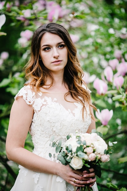 Jolie mariée avec un bouquet de mariage debout sur fond de fleurs violettes de magnolia et de verts. la nature. en plein air. regardant de côté. demi-longueur supérieure.