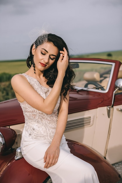 Jolie mariée élégante assise sur une voiture rétro rouge sur une route de campagne