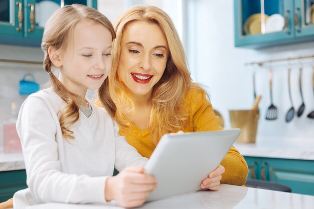 Jolie mère mince blonde souriante et à l'aide d'une tablette avec sa fille assise à la table