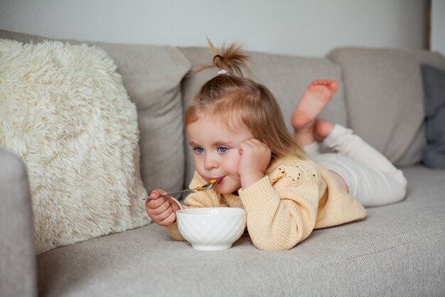 Une jolie petite fille de 2 ans dans un pull en tricot confortable prend son petit déjeuner sur le canapé