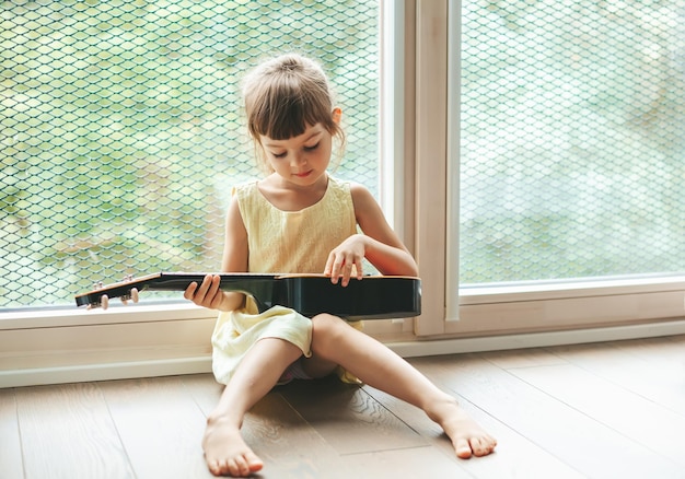 Jolie petite fille de 4 à 5 ans apprenant à jouer du ukulélé, assise sur le sol près de la grande fenêtre