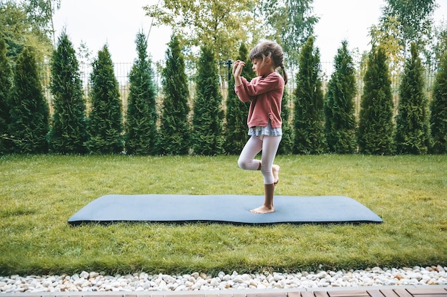 Jolie petite fille de 56 ans debout dans la position de l'Aigle ou Garudasana tout en pratiquant le yoga en plein air Yoga pour enfants concept