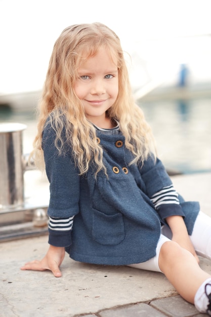 Jolie Petite Fille De 6 à 7 Ans Assise Sur Le Front De Mer à L'extérieur. Enfant  Enfant Souriant. Regarder La Caméra.