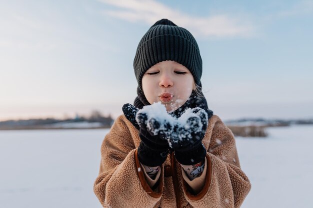 Une jolie petite fille de 6 ans souffle des flocons de neige de ses mains dans la direction de la caméra qui la photographie