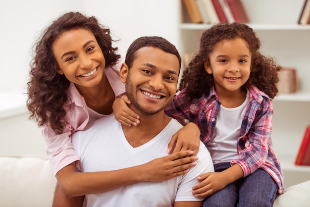 Jolie petite fille afro-américaine et ses beaux jeunes parents s'embrassant, regardant la caméra et souriant tout en étant assis sur un canapé dans la pièce.