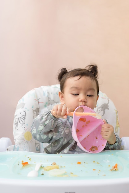 Une jolie petite fille asiatique assise sur une table à manger s'entraîne à utiliser une cuillère pour manger de la nourriture par elle-même, concept de sevrage dirigé par bébé
