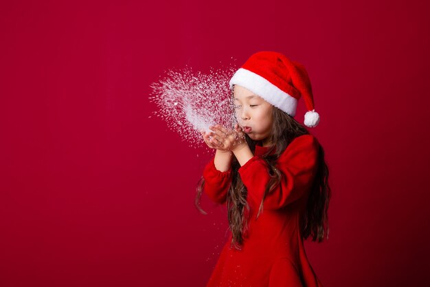 jolie petite fille asiatique dans un bonnet de noel souffle la neige de ses paumes sur fond rouge